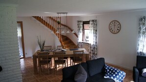 A dining area with a wooden table and chairs, a staircase, and a living room with a sofa and a clock on the wall.
