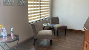 A room with a wooden floor, two grey armchairs, a small glass table with a book, and a window with blinds. - Casagrande Hotel (Monclova)
