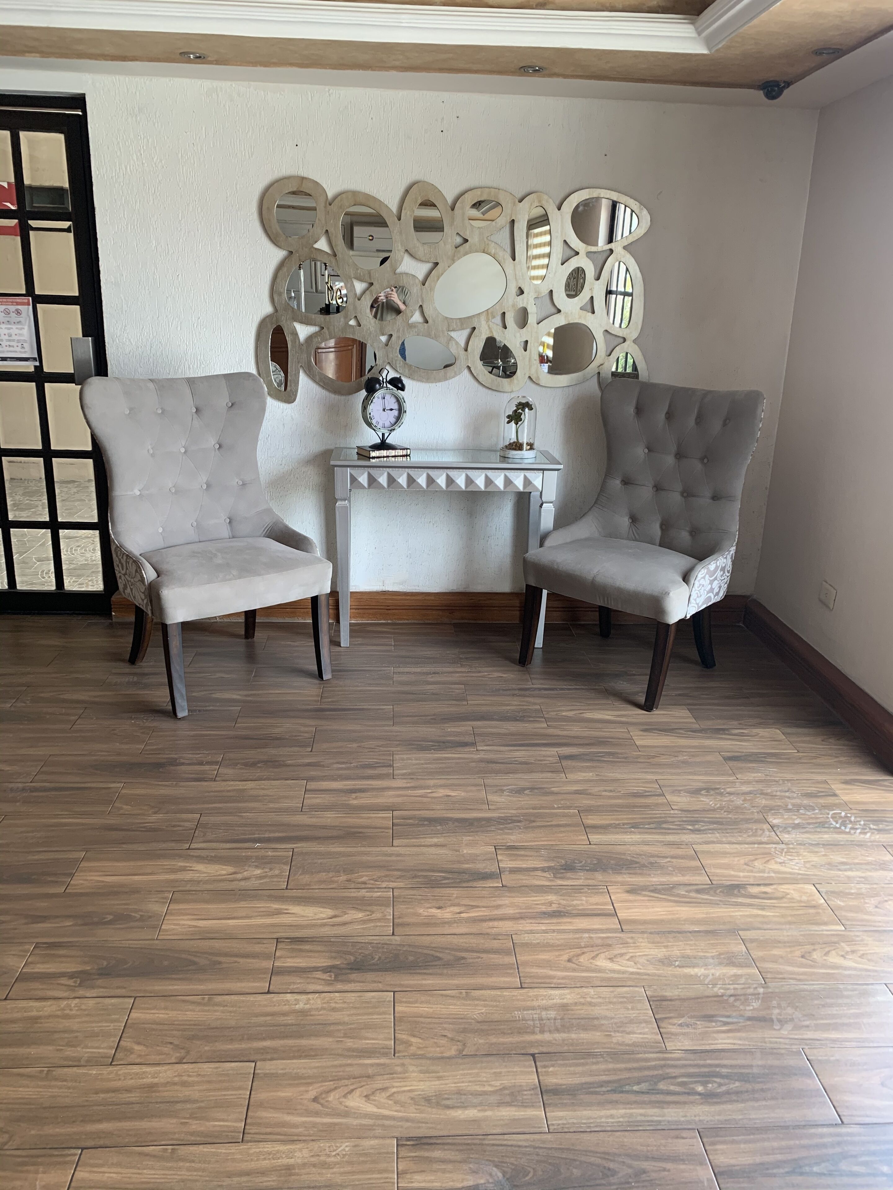 A room with two grey upholstered armchairs, a small white console table with a clock and decorative items, and a wooden herringbone floor.