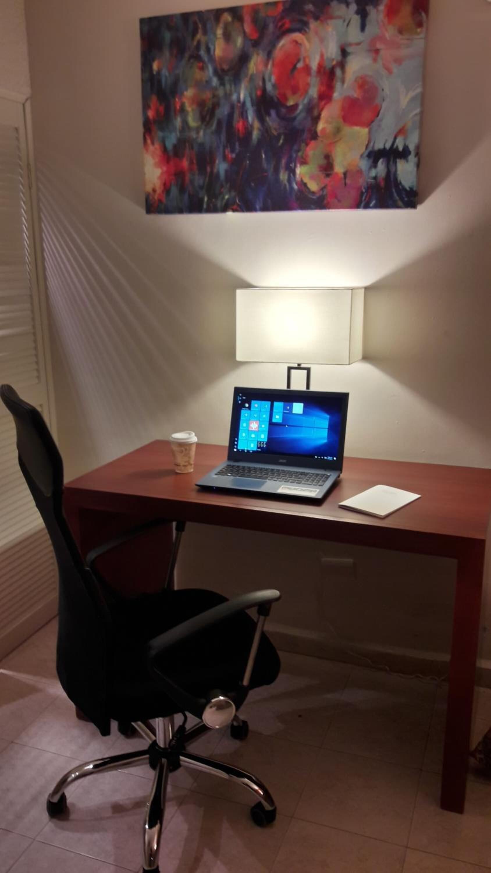 A hotel workspace with a laptop on a desk, a black office chair, a coffee cup, and a painting on the wall.