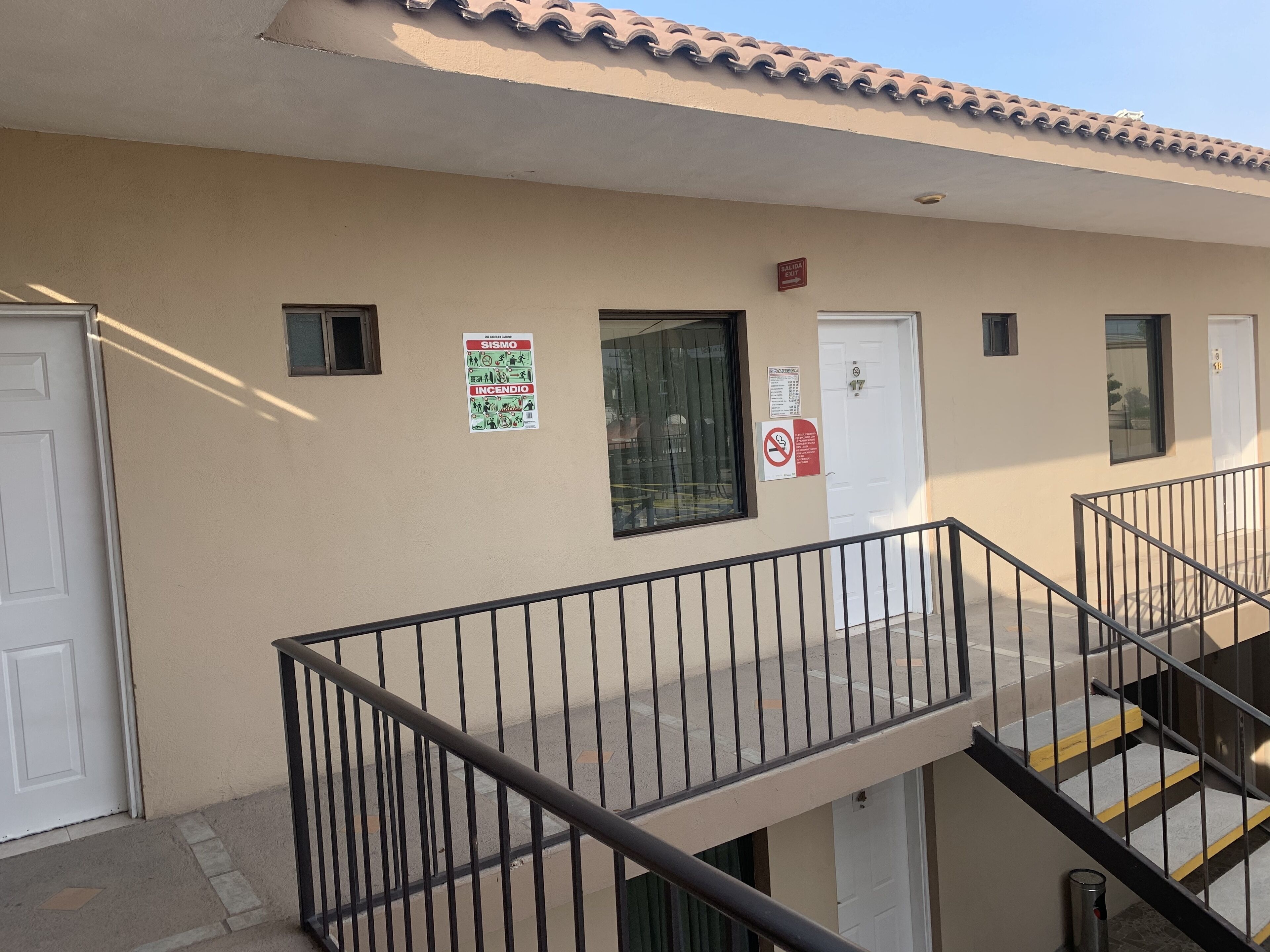 Fachada de un edificio con pared beige, techo de tejas y barandilla de metal. Se observan dos puertas, una con un letrero que dice 'SALIDA' y otra con la indicación 'No Smoking'.