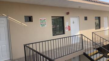 Fachada de un edificio con pared beige, techo de tejas y barandilla de metal. Se observan dos puertas, una con un letrero que dice 'SALIDA' y otra con la indicación 'No Smoking'.