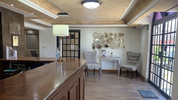 A reception area with a wooden reception desk, two armchairs, a small table with decorative items, and a wall-mounted mirror. - Casagrande Hotel (Monclova)