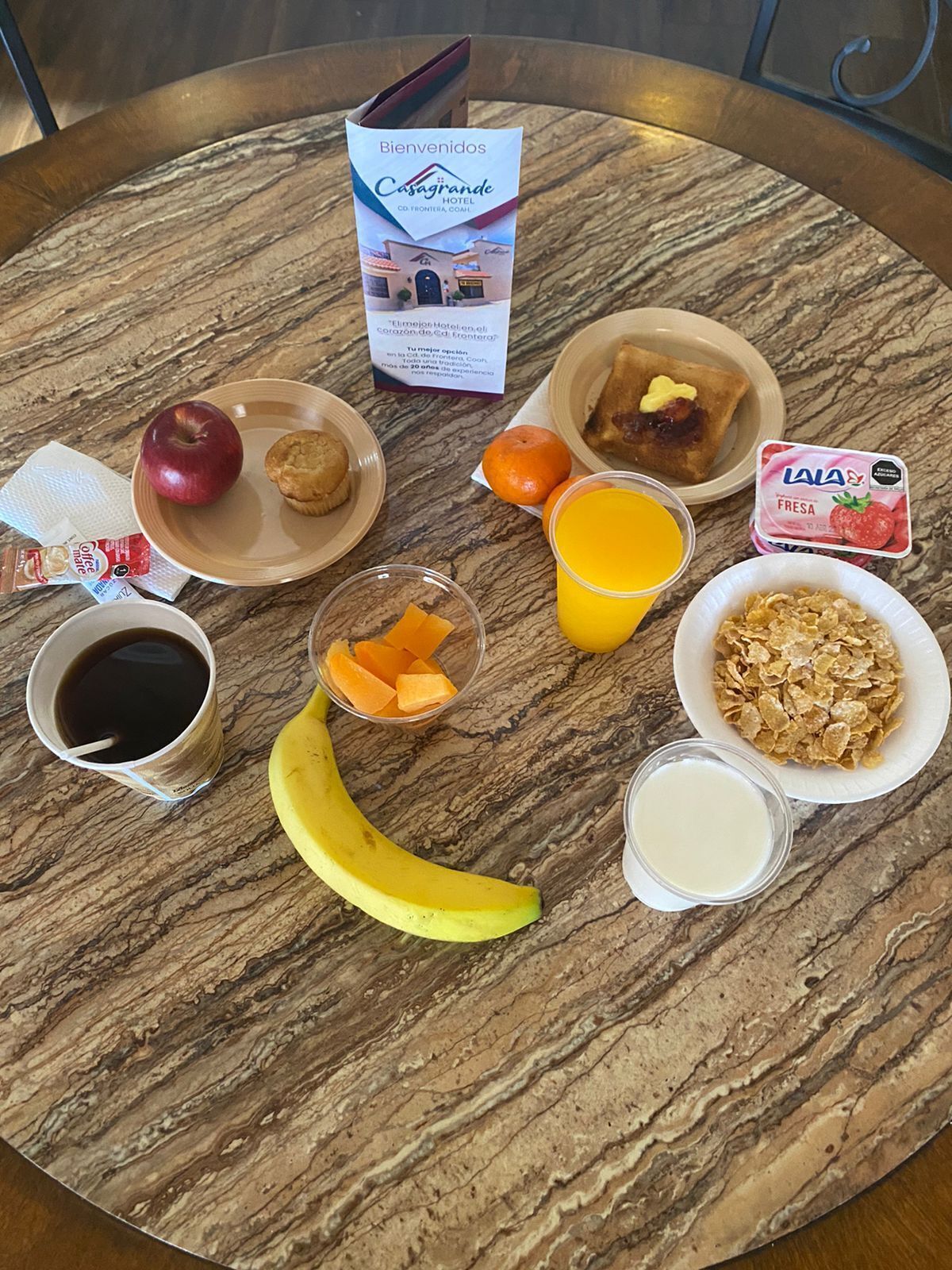 A breakfast table with a menu, apple, muffin, orange, banana, juice, cereal, milk, and a coffee cup.