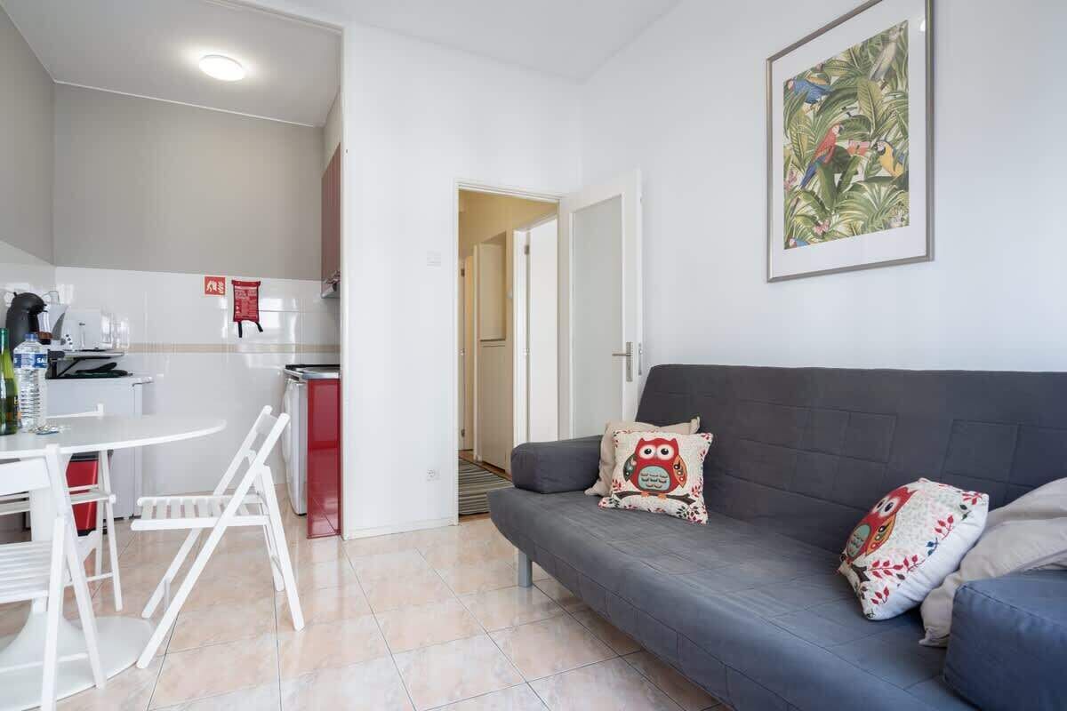 Ein modernes Wohnzimmer mit einem grauen Sofa, einem weißen Esstisch mit Stühlen und einer Küche im Hintergrund.