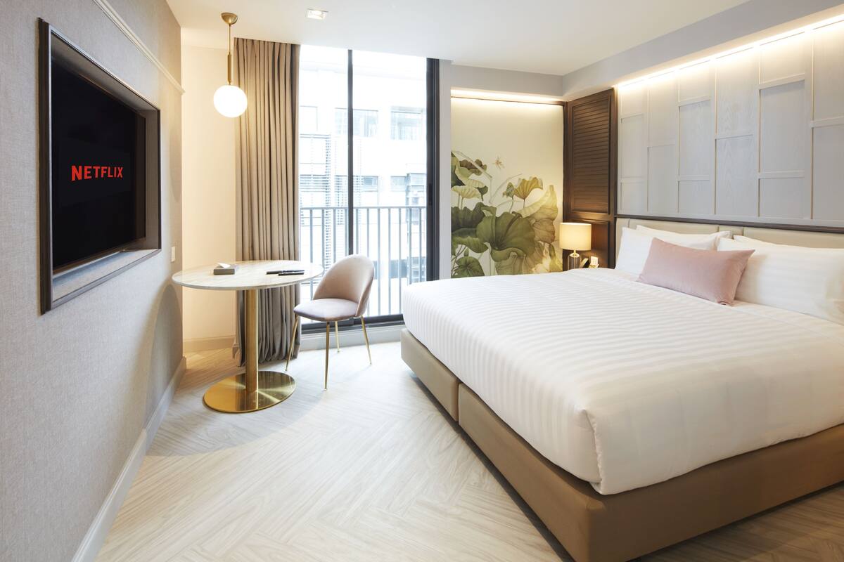 A hotel room with a large bed, a desk with a chair, a TV displaying 'NETFLIX', and a large window with a view of buildings.