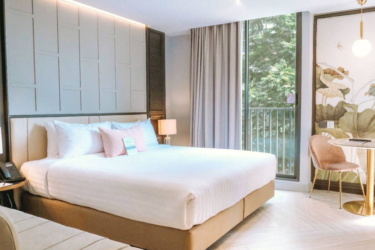 A hotel room with a large bed, a desk with a chair, a window with a view of greenery, and a wall with a floral mural.