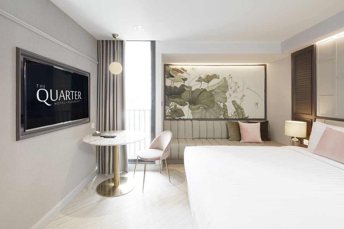 A modern hotel room with a large bed, a desk with a chair, a TV mounted on the wall, and a large floral artwork above the bed.