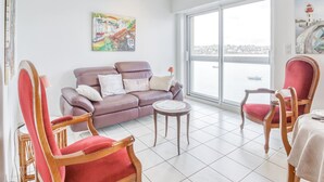A modern living room with a purple sofa, a red chair, a small round table, and a view of the city through large windows. - LE MATELOT 3* - Superbe appartement vue mer - Erquy (Erquy)