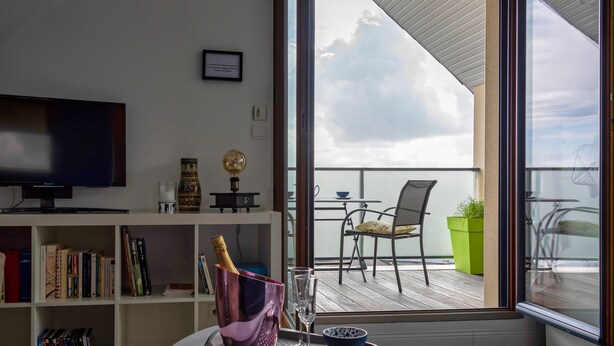 A modern living room with a flat-screen TV, a bookshelf, a small table with a tray, a champagne bottle, and a glass. A balcony with a chair and a potted plant is visible through a large window. - L'Abri du Bon Vent - Proche plage - Cancale (Cancale)