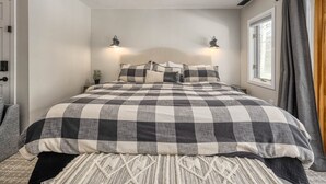 A bedroom with a checkered bedspread, two wall-mounted lamps, a window with curtains, and a door with a handle. - Ski-in/Out 1st on Mountain Lux Suite + Kitchenette (Bretton Woods)