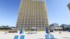 A tall hotel with multiple balconies, a beach area with lounge chairs and umbrellas, and a clear blue sky.