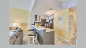 A compact apartment with a kitchenette, a living area with a sofa and bar stools, and a door leading to another room.