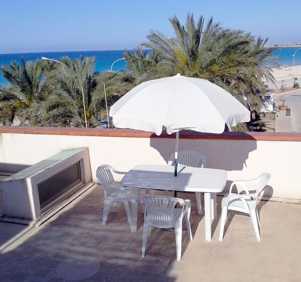 Eine Terrassengruppe im Freien mit Tisch, Stühlen, einem weißen Sonnenschirm und Blick auf das Meer und Palmen.