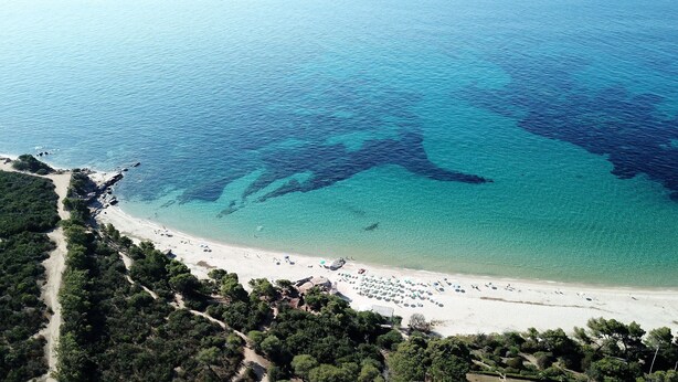 Vue aérienne d’une plage aux eaux turquoise et limpides, bordée d’un rivage sablonneux et parsemée de quelques zones de végétation.