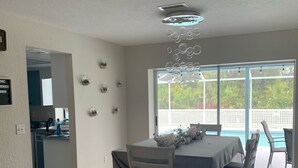 A dining area with a table and chairs, a ceiling light fixture, and a view of a swimming pool through large windows. - Heated pool for Your next vacation getaway! (North Port)