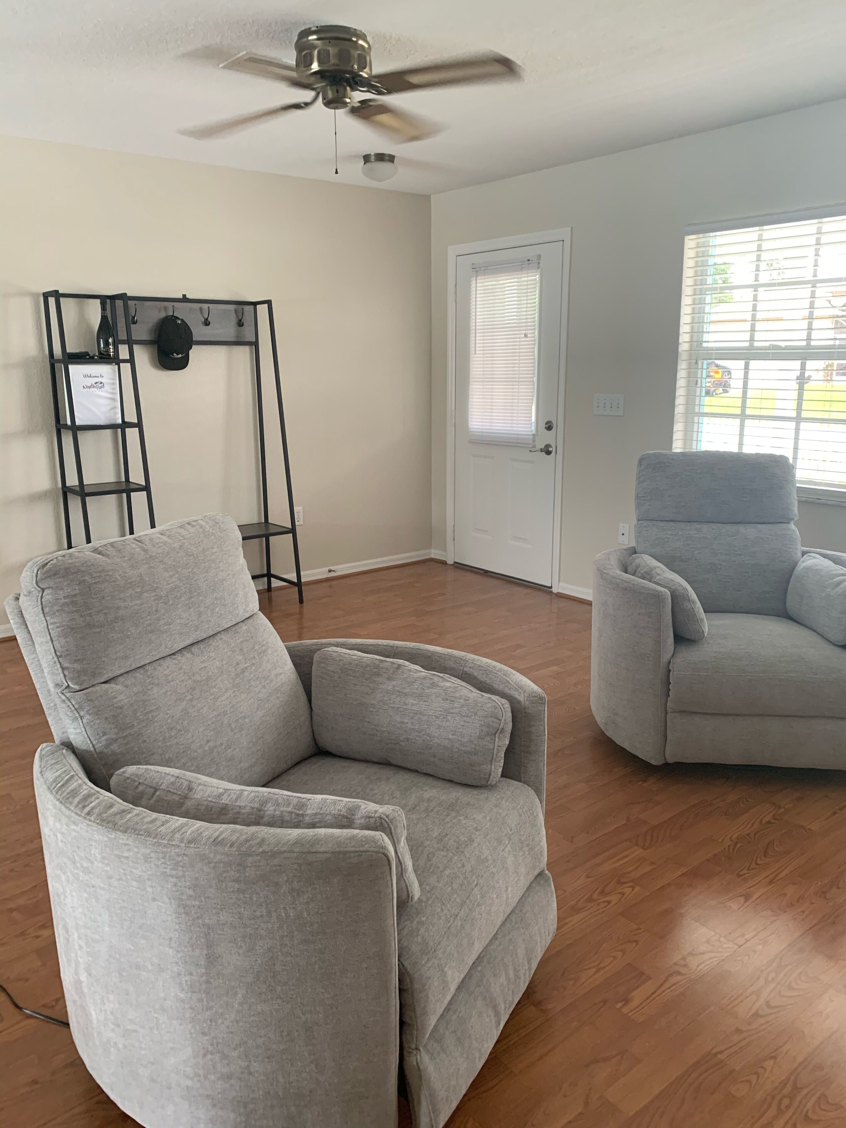 Ein Zimmer mit zwei grauen Sesseln, einem Deckenventilator, einem Kleiderständer mit Hut und Tasche, einer Tür mit Jalousien und einem Fenster mit Jalousien.