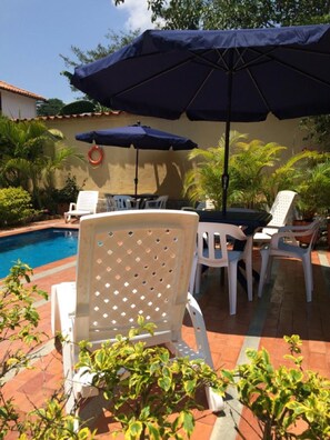 A poolside area with white plastic chairs and a large dark umbrella providing shade. - La Buganvilla Barichara (Barichara)