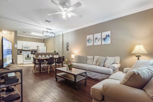 A modern living room with a sofa, coffee table, and TV, connected to a kitchen with white cabinets and appliances. - 3 BD/ 2 BA/ Lucaya Village (1-102) (Kissimmee)