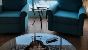 Una habitación con dos sillones color turquesa frente a una mesa de vidrio con un control remoto y una cesta de conchas marinas. Un ventanal grande ofrece vistas a una piscina y una playa.