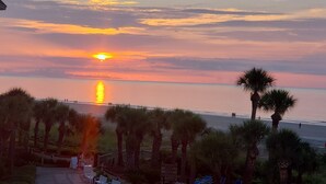 Un atardecer sobre una playa con palmeras y un paseo marítimo.