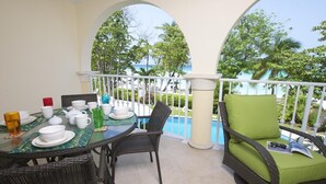 Un balcon avec une table à manger pour deux, une chaise en osier avec un coussin vert, et une vue sur l’océan et les palmiers.