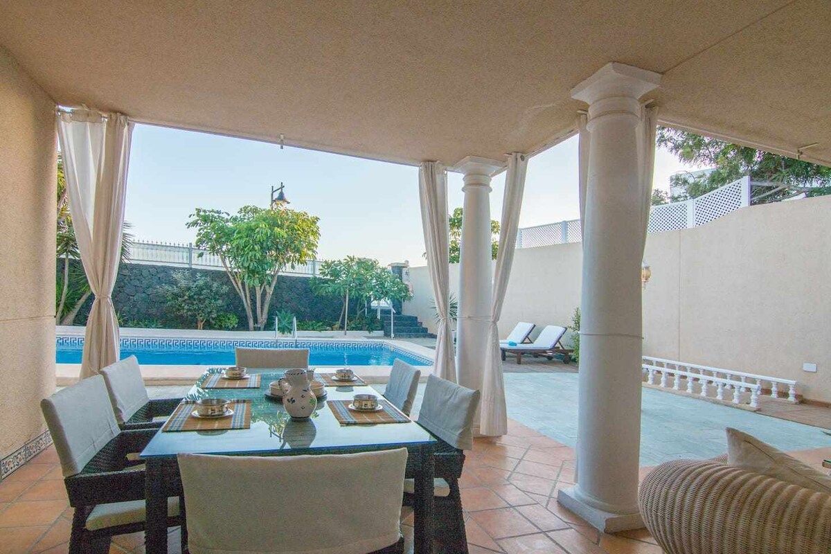 Zona pranzo all'aperto con vista piscina, coperta da un tettoia, arredata con tavoli e sedie.