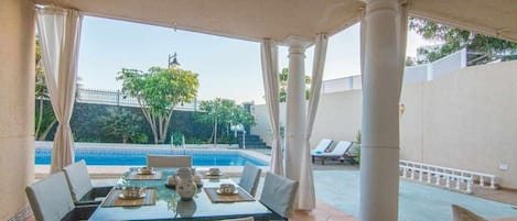 Zona pranzo all'aperto con vista piscina, coperta da un tettoia, arredata con tavoli e sedie.