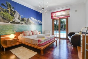 A bedroom with a bed, a nightstand, a lamp, a large mural of a beach scene, and a view of the outdoors.