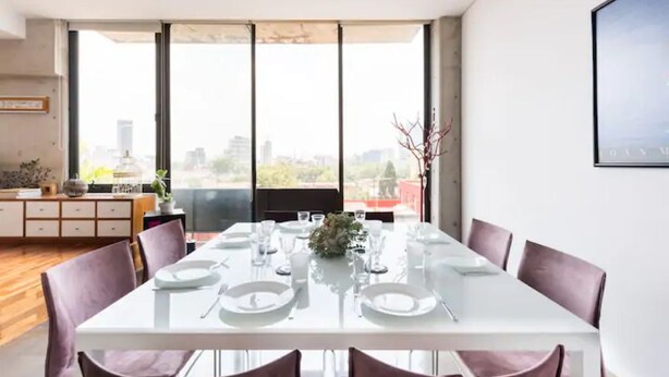Un espace de repas moderne avec une table blanche dressée pour quatre personnes, de grandes fenêtres offrant une vue sur la ville et un décor minimaliste.
