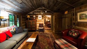 Een rustiek interieur van een blokhut met een houten plafond, een bank, een rode leren fauteuil, een bijzettafeltje en een kamerkleed met dessin.