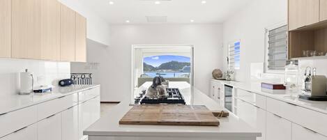 A modern kitchen with white cabinetry, a central island with a kettle, and a view of the sea through an open door.