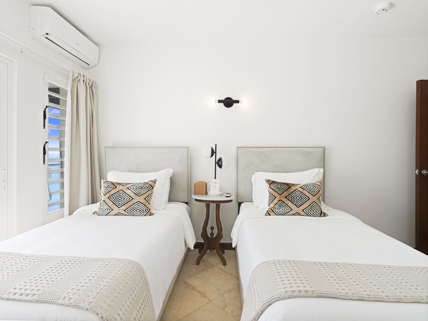 A hotel room with two beds, a small bedside table, a nightstand, a wall-mounted air conditioner, and a window with curtains.