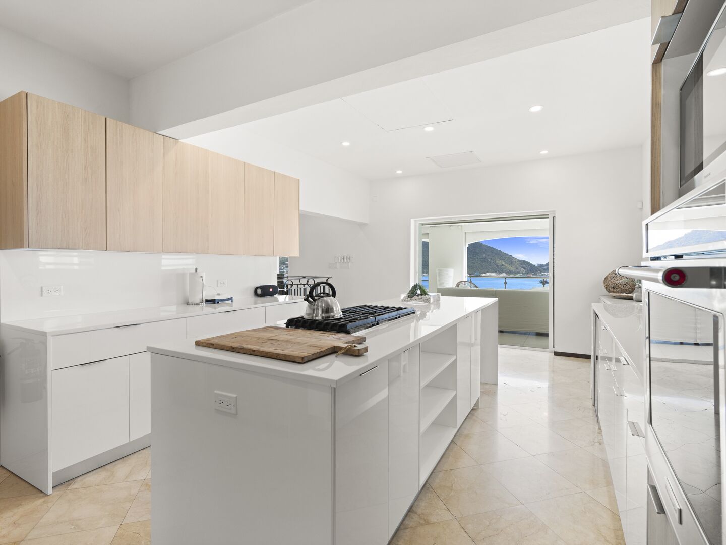 Una cocina moderna con armarios blancos, una isla central y vistas al mar a través de un amplio ventanal.