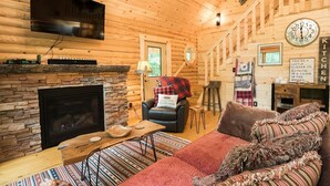 A cozy cabin interior with a stone fireplace, a television mounted on the wall, a kitchen area with a clock, and a comfortable seating area with a couch and chairs. - Secluded, pet-friendly, new log cabin. (Morgantown)