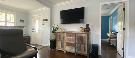 A spacious living room with hardwood flooring, a large flat-screen TV mounted on the wall, a wooden cabinet with decorative items, and a comfortable chair.