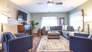 A living room with a gray sofa, two armchairs, a coffee table, a patterned rug, a ceiling fan, a flat-screen TV, a fireplace, and a window with sheer curtains. - Pet-friendly, renovated, walk to downtown. (Nashville)