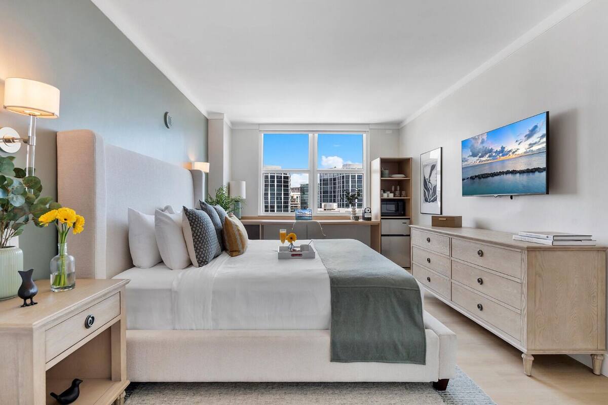 A modern bedroom with a large bed, bedside tables, a TV, a window with a city view, and a desk with a chair.