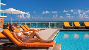 A poolside area with lounge chairs and umbrellas, a clear sky, and a view of the ocean. - Private Oasis at Arya (Miami)