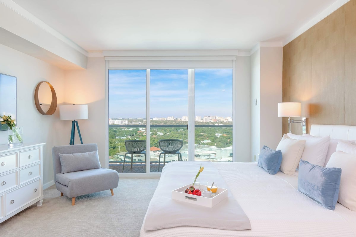 A hotel room with a large bed, a chair, a dresser, and a view of the city through large windows.