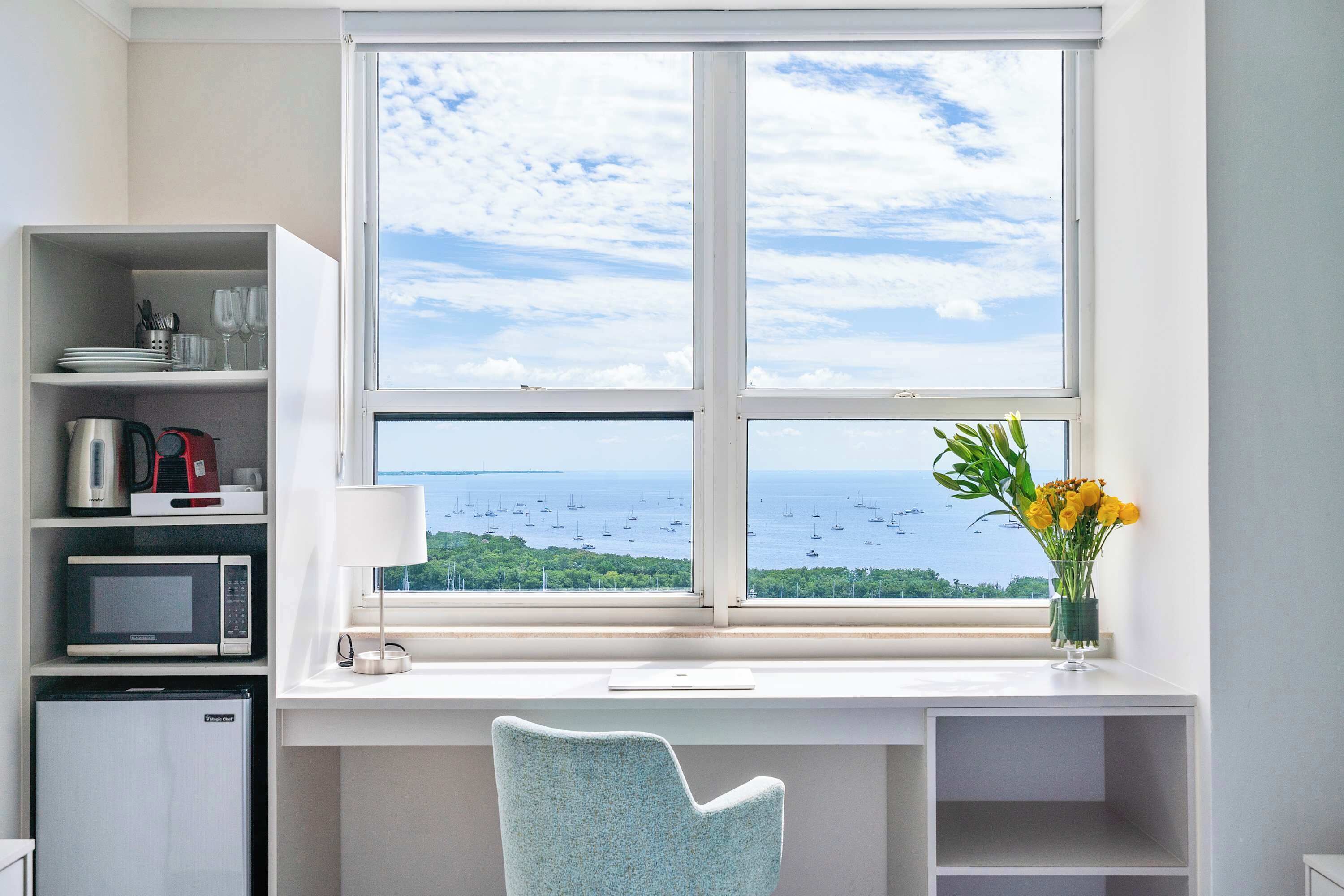 A room with a desk, a chair, a microwave, a refrigerator, a lamp, and a window with a view of the sea and boats.