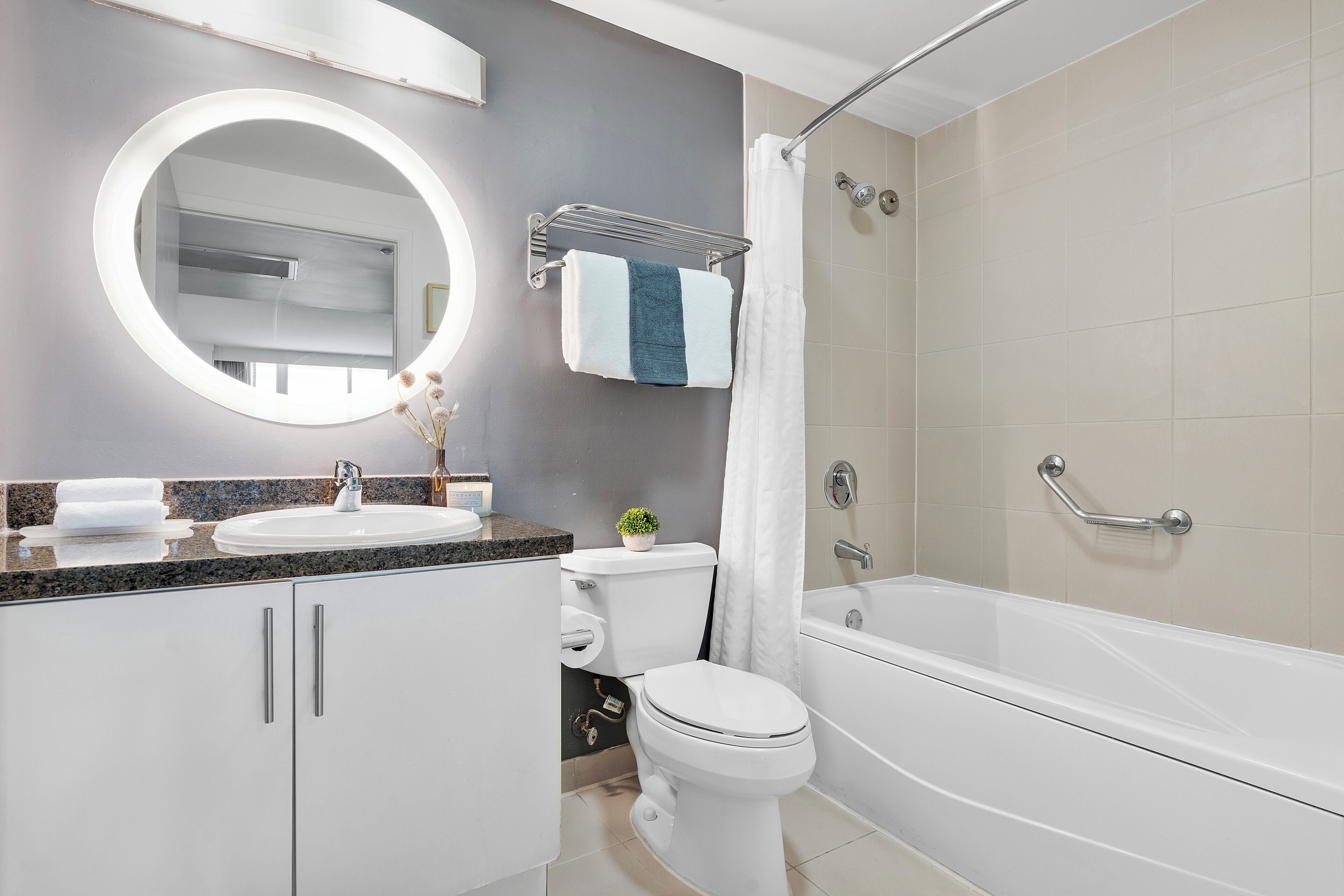 A modern bathroom with a white sink, a round mirror with lights, a toilet, and a bathtub.