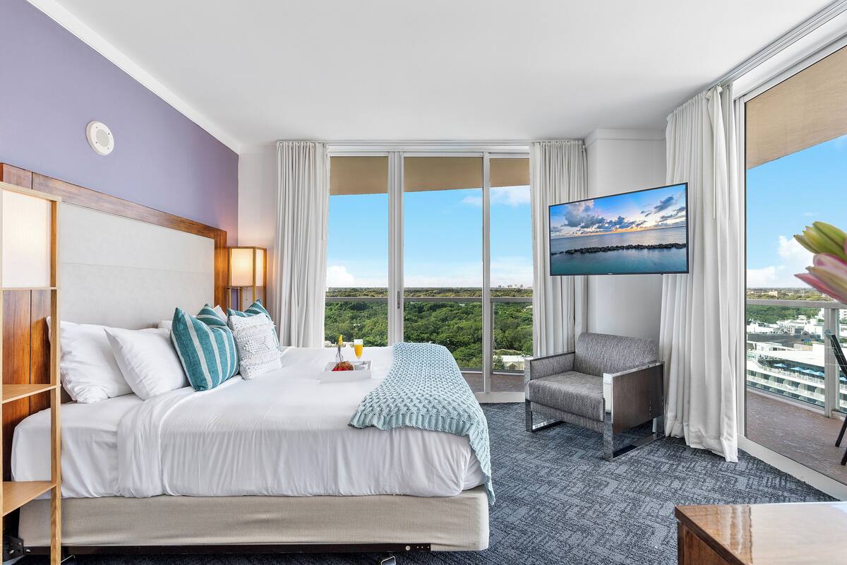 A hotel room with a large bed, a TV, a balcony with a city view, and a desk.
