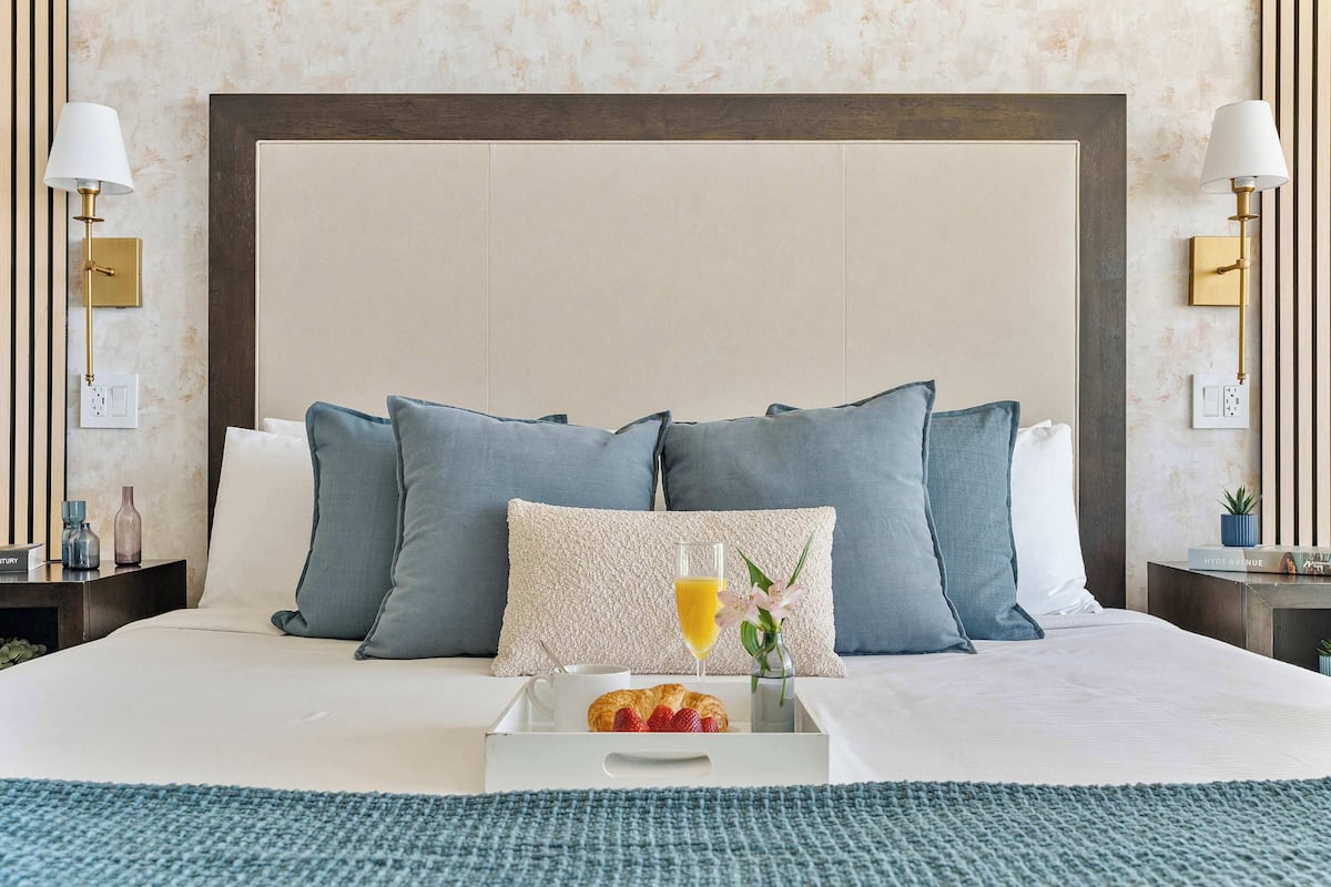 A neatly made bed with pillows, a tray with croissants and a drink, and a bedside table with a lamp and a small plant.