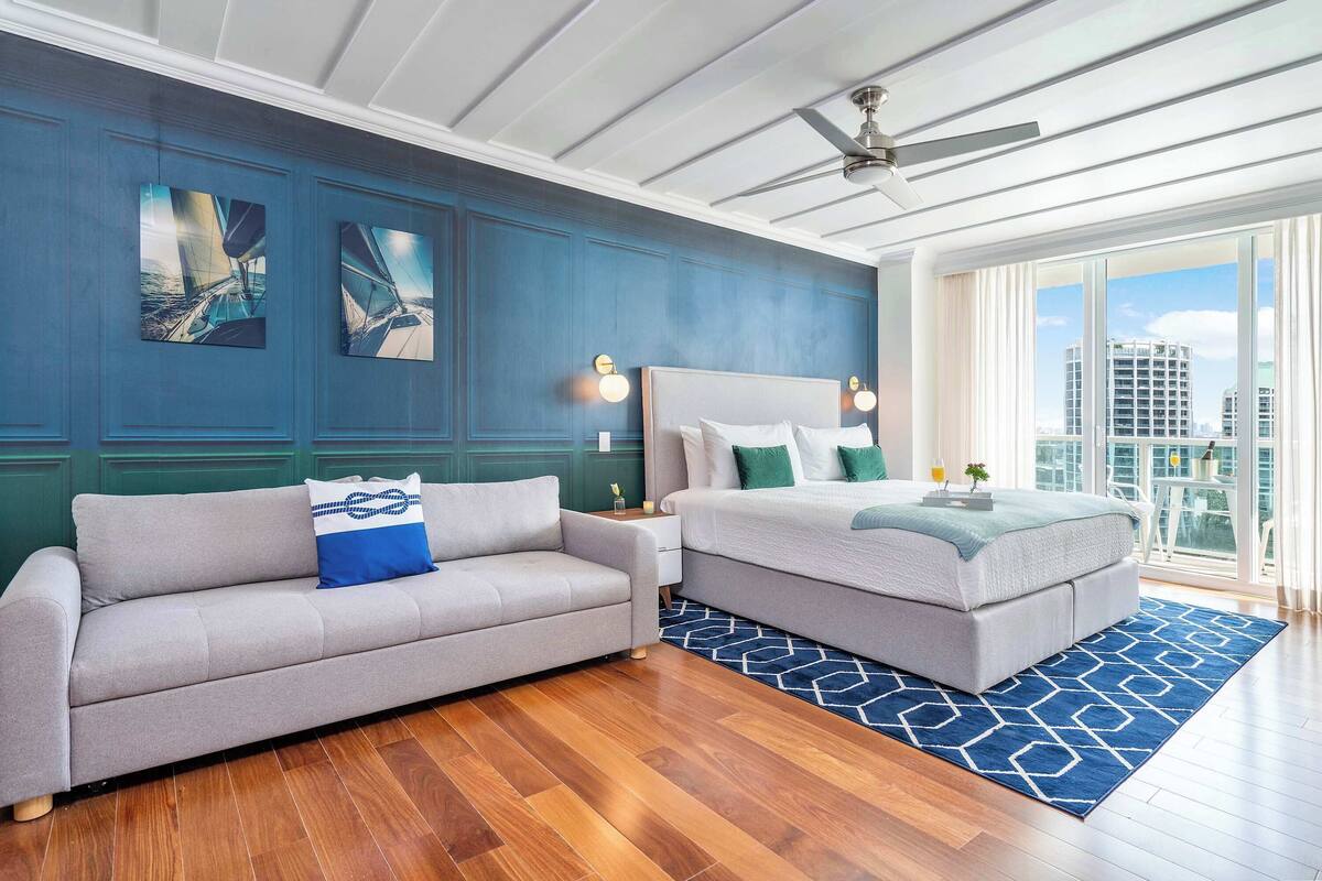 A modern hotel room with a sofa, a bed, a ceiling fan, and a view of the city.