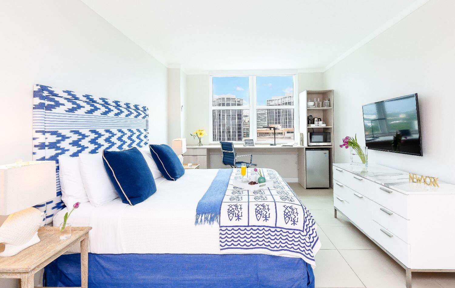 Une chambre d’hôtel moderne équipée d’un lit, d’un bureau, d’une télévision et offrant une vue sur les bâtiments grâce à de grandes fenêtres.