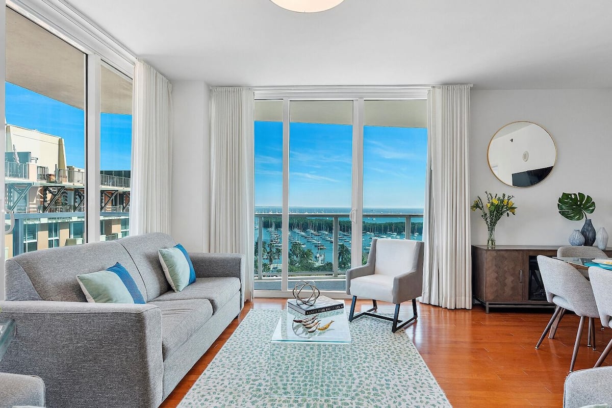 A modern living room with a grey sofa, a small table, a chair, and a view of the marina through large windows.
