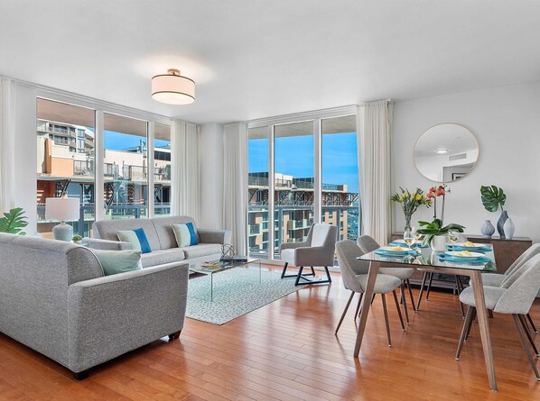 A modern living room with a sofa, dining table, chairs, and large windows offering a city view. - Private Oasis at Arya (Miami)