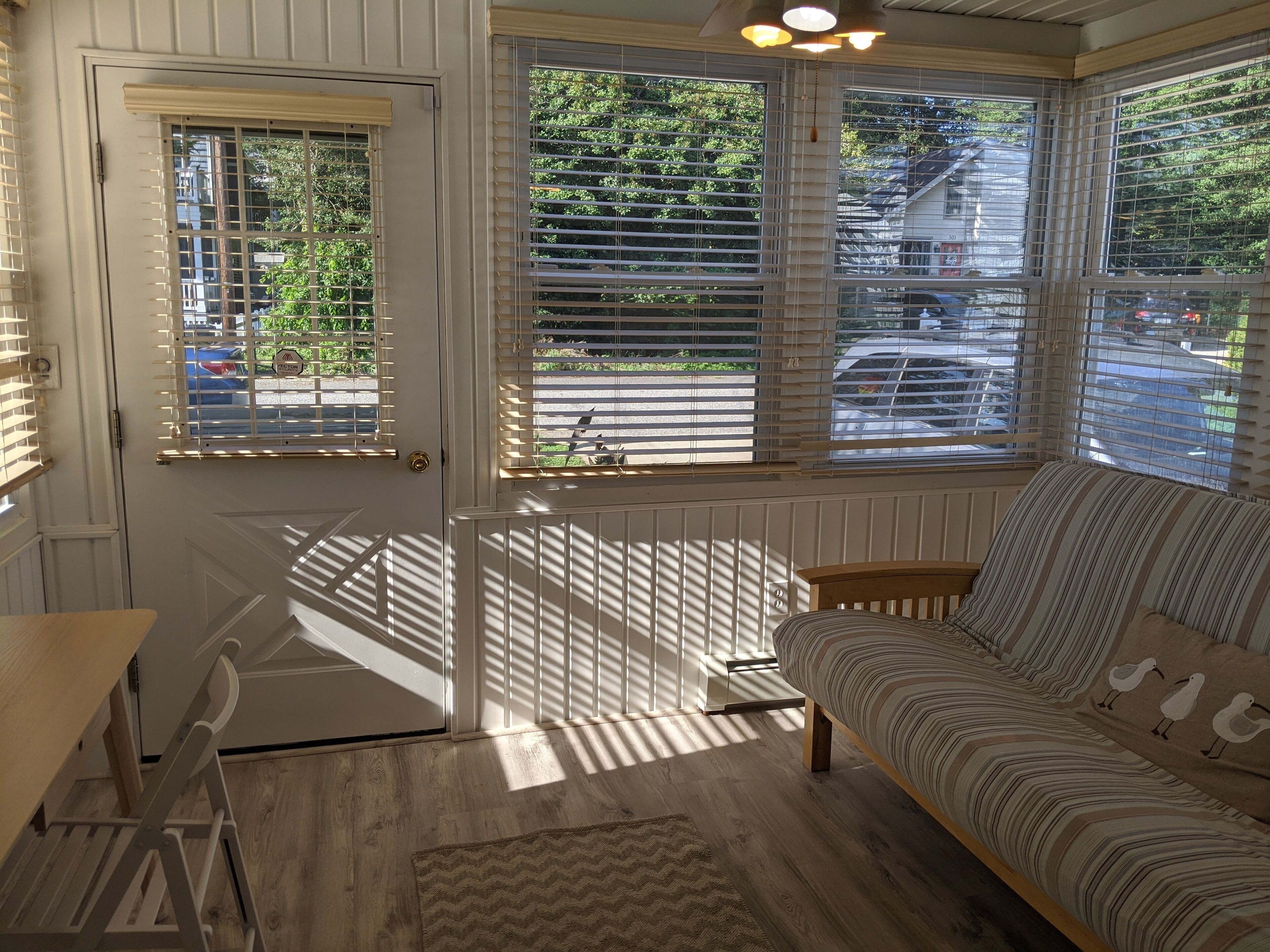 A cozy room with a wooden floor, a striped sofa, and a view of the street through large windows with blinds.