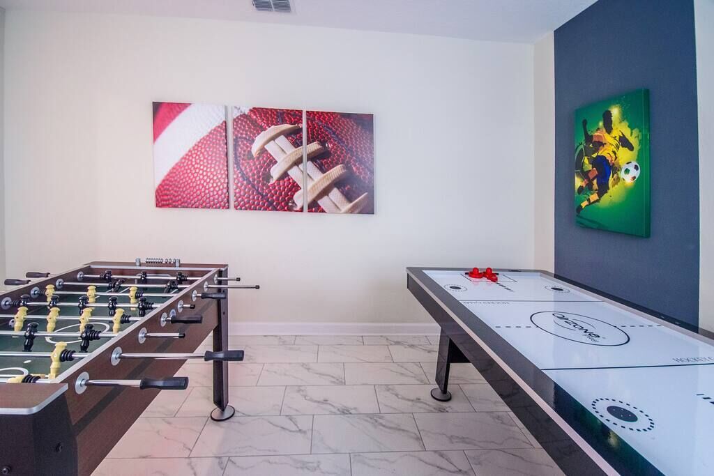 A room with a foosball table and shuffleboard table, both in neutral colors, with a simple tiled floor and plain walls.